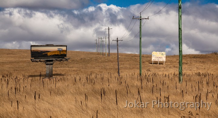 Cooma Welcomes You.jpg - Cooma wlecomes you.  The road down through the Monaro towards Cooma is lined with innumerable advertising billboards, navigational signposts and ‘Welcome to…’ signs. My favourite is a sign which simply declares: “Monaro Merino Country”. It’s straightforward, unequivocal, indeed perhaps a little blunt. This is sheep country.The passing motorist can have no doubt about how to regard the landscape that expands before them as they head south - the gold and brown treeless plains and the undulating wooded hills, with here and there a view to the more distant ridges and peaks of the Great Dividing Range. Alternate readings of the land are not countenanced - or even acknowledged.A little further south, and on the other side of the road, beside a long straight line of old power transmission poles, is another sign to warmly advise all and sundry that “Cooma Welcomes You”. Similar signs can be found on the outskirts of most towns, and are probably a world-wide phenomenon. (“Pyongyang Welcomes You”?) But I’ve always been puzzled by these all-encompassing, anonymous messages of civic goodwill, because I’m sure that there are people who are NOT welcome in Cooma (or other places).In this image, I’ve inserted another, more enigmatic sign, with the slightly cryptic assertion (as a billboard within the billboard) that this is in fact ‘Vacant Land’. All claims on the land, all claims of possession, control or exclusive usage are rejected, as this is Terra Nullius.  (But if this were indeed the case, who would have the authority to erect such a sign?!)